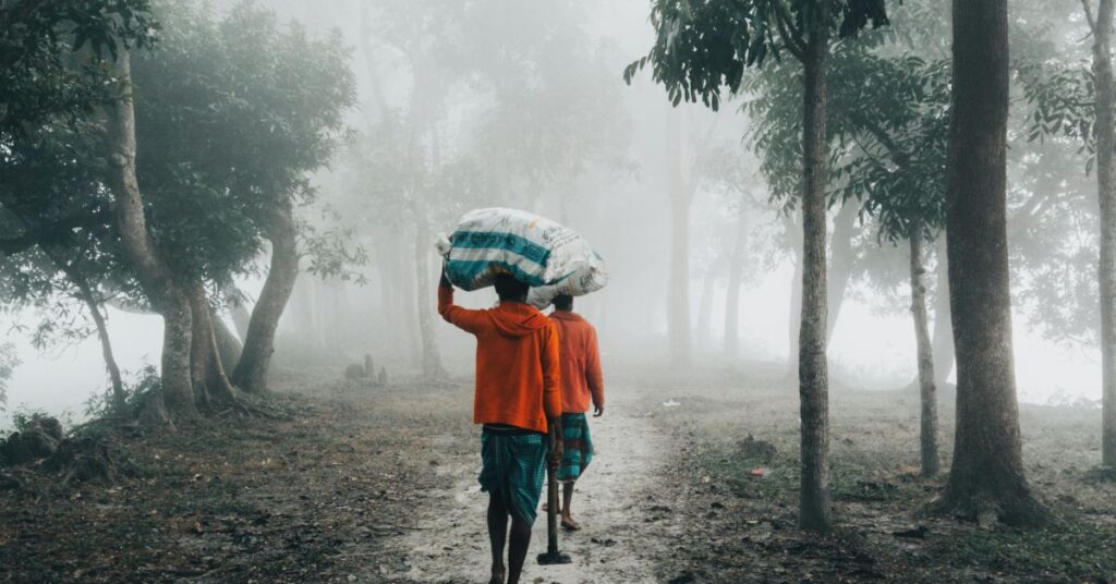पेड़ों से घिरे धुंधले रास्ते पर कंधे पर बोझ लिए चलते किसान, भारतीय गाँव की सादगी और श्रम की झलक दिखाता दृश्य।