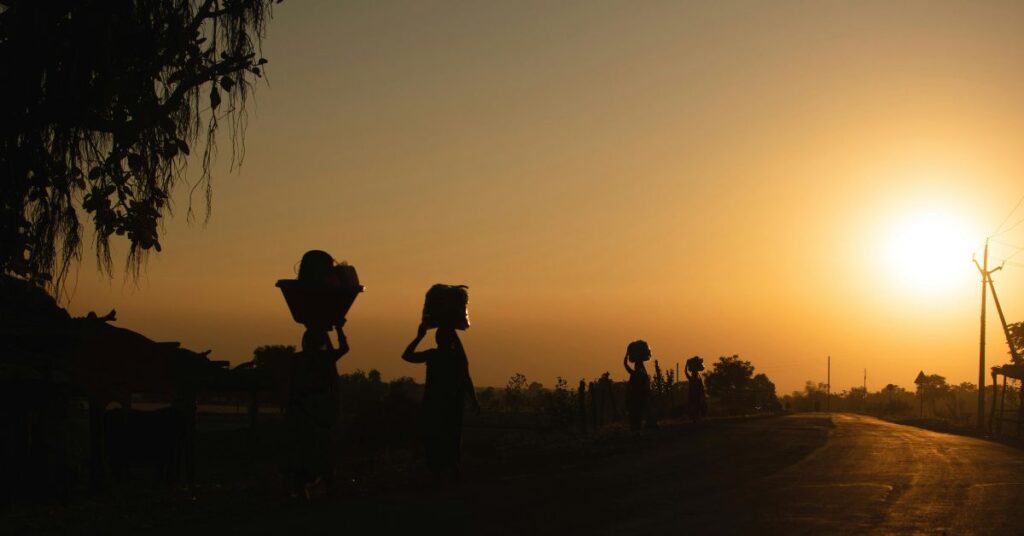 सूर्यास्त में लौटता हुआ गाँव का जीवन” सूर्यास्त के समय गाँव की सड़क पर सिर पर सामान लिए जाती महिलाएँ, भारतीय ग्रामीण जीवन का सादगी भरा दृश्य।