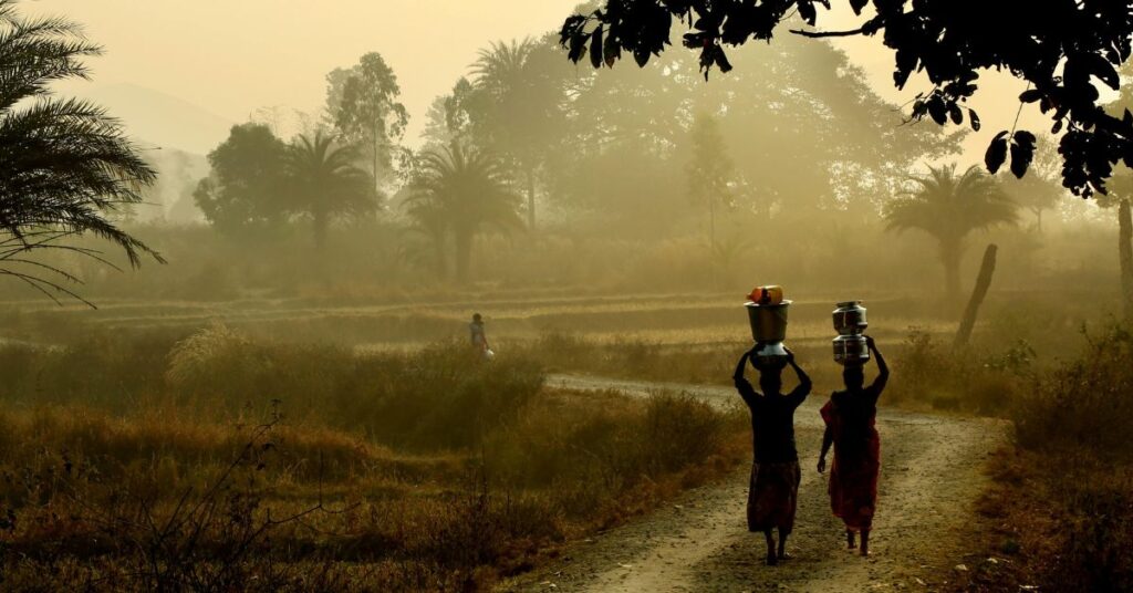 भोर की रोशनी में गाँव की दिनचर्या सुबह के समय खेतों के रास्ते पर सिर पर बर्तन लिए जाती ग्रामीण महिलाएँ, भारतीय गाँव की शांत दिनचर्या को दर्शाता चित्र।